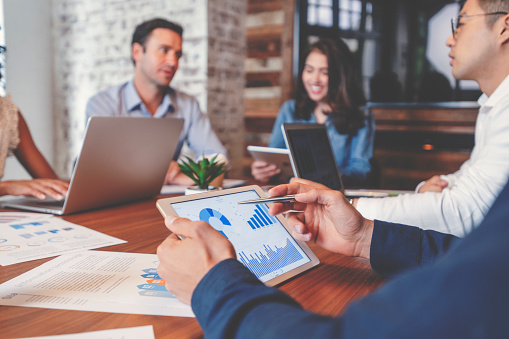 Group of people meeting with technology. There are marketing, financial and strategy documents on the wooden board room table and on the computers and digital tablets. Multi ethnic group including Caucasian and African American. 